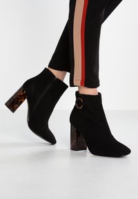 Botines de ante negros con diseño de punta afilada, que cuentan con un tacón en bloque de carey y una cremallera lateral. Acentuados con un detalle de hebilla circular.