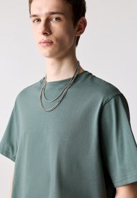 Groene katoenen t-shirt met korte mouwen en een ronde hals, met een gladde textuur. Gelaagde zilveren kettingen voegen detail toe.