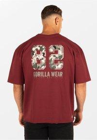 Bordeauxkleurig t-shirt met korte mouwen en grote camouflage cijfers "82" op de achterkant, met daaronder de tekst "GORILLA WEAR". Zacht, casual materiaal.