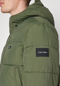 Mand iført en olivengrøn polstret jakke med hætte og et sort Calvin Klein-logo patch på den øverste ærme.