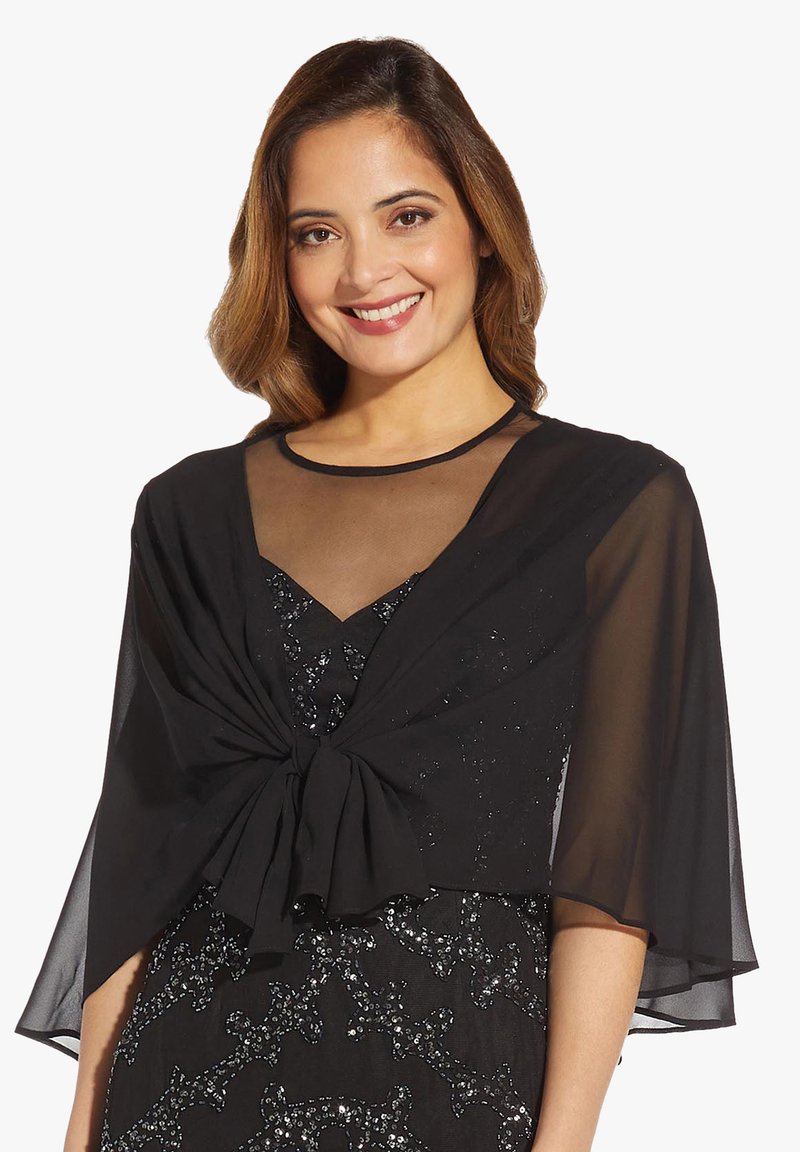 Donna sorridente che indossa un scialle trasparente nero legato sopra un abito nero con paillettes e scollatura in rete.