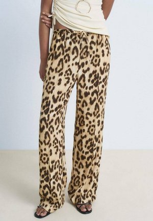 Persoon draagt beige wijde broek met luipaardprint, wit gedrapeerd bovenstuk met grote hoepel detail en zwarte sandalen met open tenen.