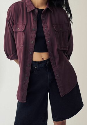 Personne portant une chemise bordeaux ouverte sur un top court noir et un short taille haute noir, debout devant un fond uni.