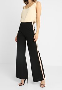 Pantaloni neri a gamba larga con una striscia laterale rosa, abbinati a un top beige senza maniche. Presentano un tessuto morbido e una vestibilità rilassata.