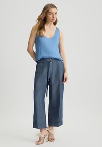 Modrá bezrukávová blúzka skombinovaná s voľnými nohavicami pripomínajúcimi denim a béžovými sandálmi na vysokom opätku s pierkovými akcentmi. Ploché, minimalistické pozadie.