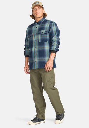 Rutet flannel skjorte i blå, grønne og orange nuancer, med to brystlommer og knaplukning, kombineret med olivengrønne bukser og sneakers.