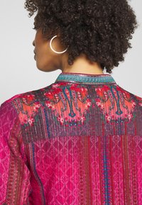 Farbiger, gemusterter Stoffoberteil in rosa und roten Farbtönen, mit floralen Motiven, strukturierten Streifen und einem verzierten Ausschnitt.