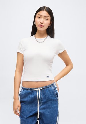 Jeune femme aux longs cheveux noirs portant un t-shirt blanc court à manches courtes et un pantalon en jean bleu ample avec cordon, debout avec une main sur la hanche.