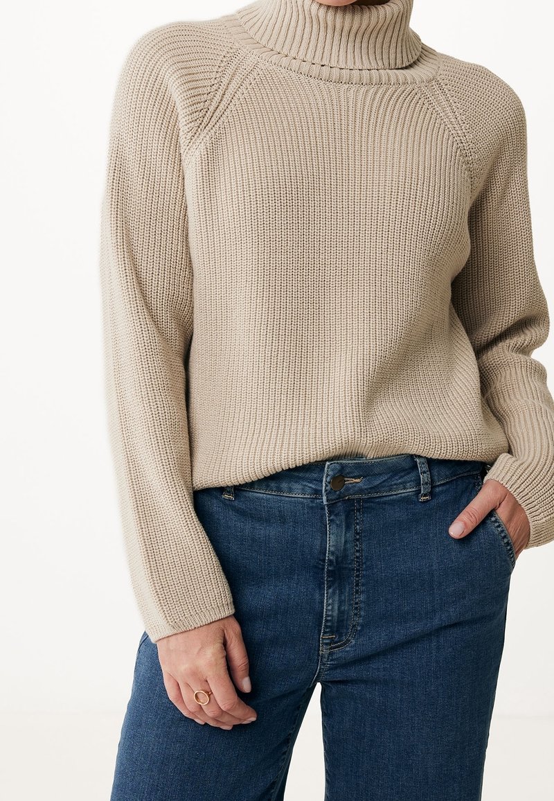 Pull à col roulé beige en tricot porté avec un jean bleu en denim, une main dans la poche et l’autre reposant sur le côté avec une bague simple.