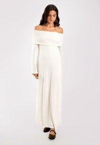 Schulterfreies, langes weißes Strickkleid mit langen Ärmeln und fließender Silhouette, ergänzt durch braune geschlossene Schuhe.