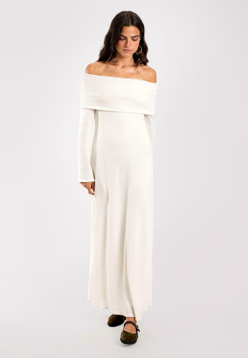 Schulterfreies, langes weißes Strickkleid mit langen Ärmeln und fließender Silhouette, ergänzt durch braune geschlossene Schuhe.