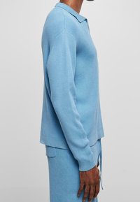 Ljusblå långärmad stickad tröja med ribbad textur, kragdesign och sidofickor i nederkant för ökad rörelsefrihet.