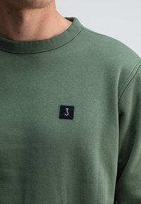 Groene sweatshirt met een ronde hals, gemaakt van zacht materiaal. Bevat een klein zwart label met een witte letter "J" op de borst.