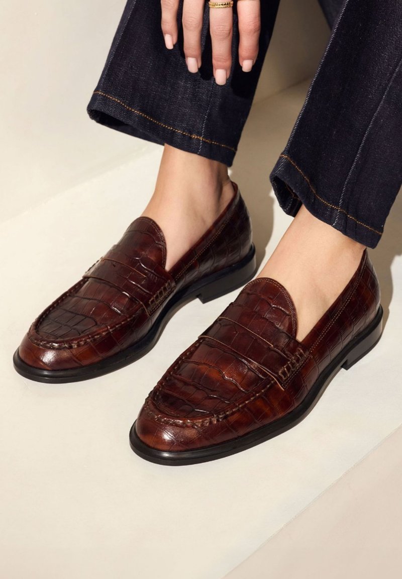 Bruine leren instappers met een krokodillenstructuur, ronde neus, decoratieve stiksels en een platte rubberen zool, gecombineerd met donkerdenim broek.