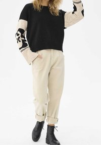 Pull en maille noire avec des manches beiges ornées d'un motif noir audacieux, assorti à un pantalon crème retroussé et des bottes noires à lacets.