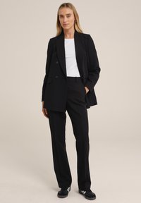 Černý dobře střižený blazer s dvouřadovým designem a klopami, doplněný o černé rovné kalhoty a bílou košili pod ním.