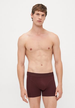 Oben ohne Mann mit kurzen braunen Haaren, der dunkelrote Boxer-Shorts trägt und gegen einen schlichten hellen Hintergrund steht, mit dem Gesicht nach vorn.