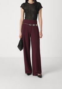 Czarna koronkowa blouse z krótkimi rękawami, zestawiona z szerokimi burgundowymi spodniami. Srebrny pasek łańcuszkowy i czarna torebka dopełniają stylizację.