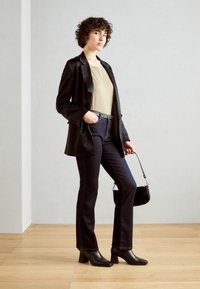 Svart kavaj över en guldtop, parat med mörka denimflaredjeans och svarta klackstövlar. Liten svart handväska och lockigt hår.