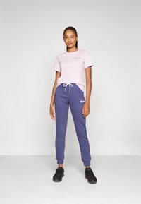 Un t-shirt en coton rose clair associé à un pantalon de jogging bleu marine avec un cordon blanc et un logo brodé. Des chaussures noires complètent la tenue.