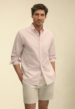 Mężczyzna z podwiniętymi rękawami, ubrany w jasnoróżową koszulę w paski na guziki oraz beżowe szorty, stoi na tle jednolitego beżowego tła.