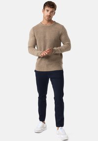 Maglione beige a maglia con texture a coste e colletto rotondo, abbinato a pantaloni blu scuro e sneakers bianche. Modella in piedi con le mani unite.