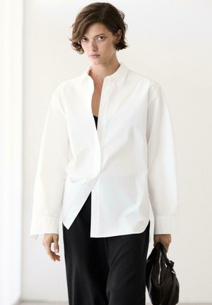 Młoda osoba z krótkimi włosami nosi oversize białą koszulę zapinaną na guziki na czarnym stroju, trzymając czarną torbę w prawej ręce.