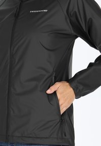 Giacca nera impermeabile con una texture liscia, chiusura frontale con zip e tasche laterali. Presenta un logo sul petto e polsini elasticizzati.