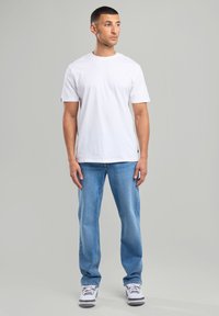 Camiseta de algodón blanca con mangas cortas, combinada con jeans de mezclilla azul claro. Los zapatos son blancos con acentos grises. Postura de pie, fondo neutro.