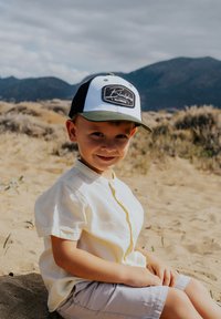 Gorra de béisbol con un diseño en blanco y negro, que presenta un parche de logo. Camisa de botones de manga corta en un amarillo claro y pantalones cortos beige. Fondo arenoso.