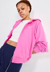 Felpa rosa con zip e logo Nike bianco, abbinata a un top corto bianco e pantaloni con cintura elastica blu navy. Tessuto morbido.