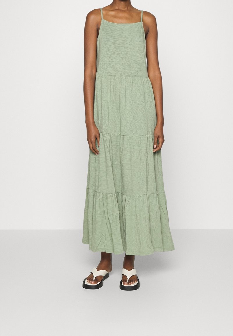 Robe maxi vert clair à plusieurs niveaux avec des bretelles spaghetti ajustables et une coupe décontractée. Portée avec des sandales plates noir et blanc.