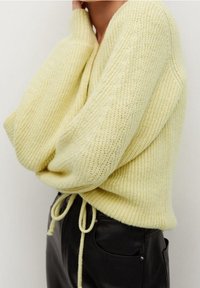 Pull jaune en maille avec manches surdimensionnées, col en V et cordon de serrage à la taille, associé à un pantalon en cuir noir.
