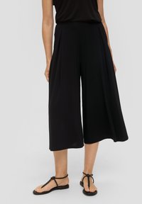 Zwarte culottes met plooien, gemaakt van een zachte stof. Kenmerkt zich door het brede broekspijpen ontwerp dat onder de knie uitsteekt, gecombineerd met platte sandalen.