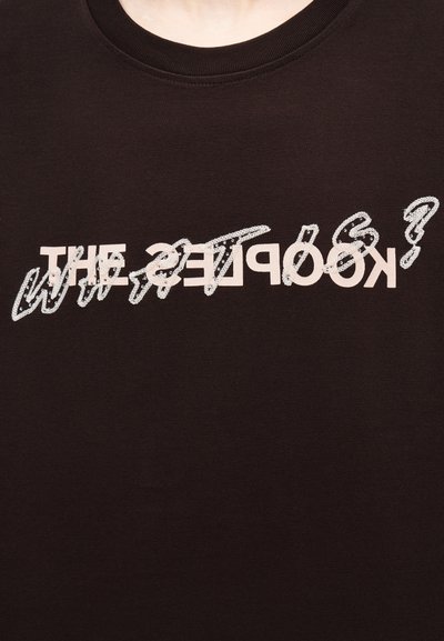 Μαύρο μπλουζάκι με λαιμόκοψη σε στυλ crew neck, με λευκό κείμενο που γράφει "THE KOOPLES?" και επάνω του, σε λευκή καλλιγραφική γραφή, η φράση "WHAT'S?" στο ύψος του στήθους.