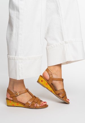 Feet wearing brown leather wedge sandals with orange polka dots on the heel, paired with white wide-leg cropped pants.