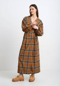 Brązowa długa sukienka w kratę z bufkami na rękawach i paskiem do wiązania. Posiada niebieskie i zielone akcenty, miękką teksturę oraz boczne rozcięcia dla swobody ruchów.