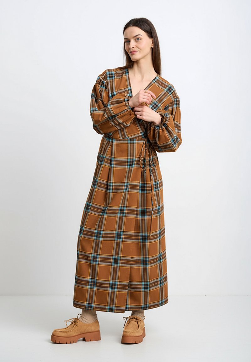 Brązowa długa sukienka w kratę z bufkami na rękawach i paskiem do wiązania. Posiada niebieskie i zielone akcenty, miękką teksturę oraz boczne rozcięcia dla swobody ruchów.