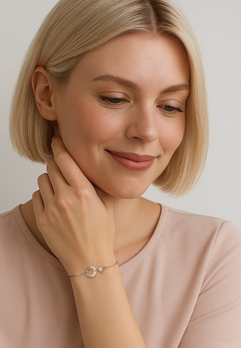Bracciale d'argento con un charm circolare e un dettaglio a forma di cuore, indossato attorno a un polso. Texture liscia contro una camicia di un rosa chiaro.