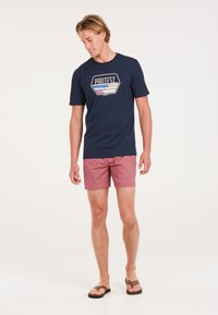 Camiseta de manga corta en color marino con un diseño de logo geométrico y shorts de baño en color coral. Los shorts cuentan con una tela texturizada y una cinturilla elástica.