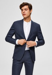 Mørkeblå dress med teksturert stoff, slim-fit design, notched lapels, og en hvit skjorte under.