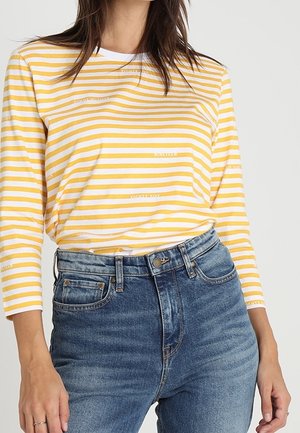 Frau trägt ein langärmeliges, gelb-weiß gestreiftes Hemd, steckt es in hoch taillierte blaue Jeans und legt die Hand seitlich ab.