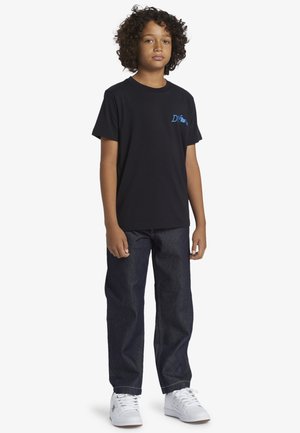 Un garçon aux cheveux bouclés portant un t-shirt graphique noir, un jean foncé et des baskets blanches, se tenant devant un fond blanc.