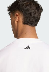 White cotton t-shirt featuring a black Adidas logo at the back, short sleeves, and a crew neckline. Soft texture and casual fit.