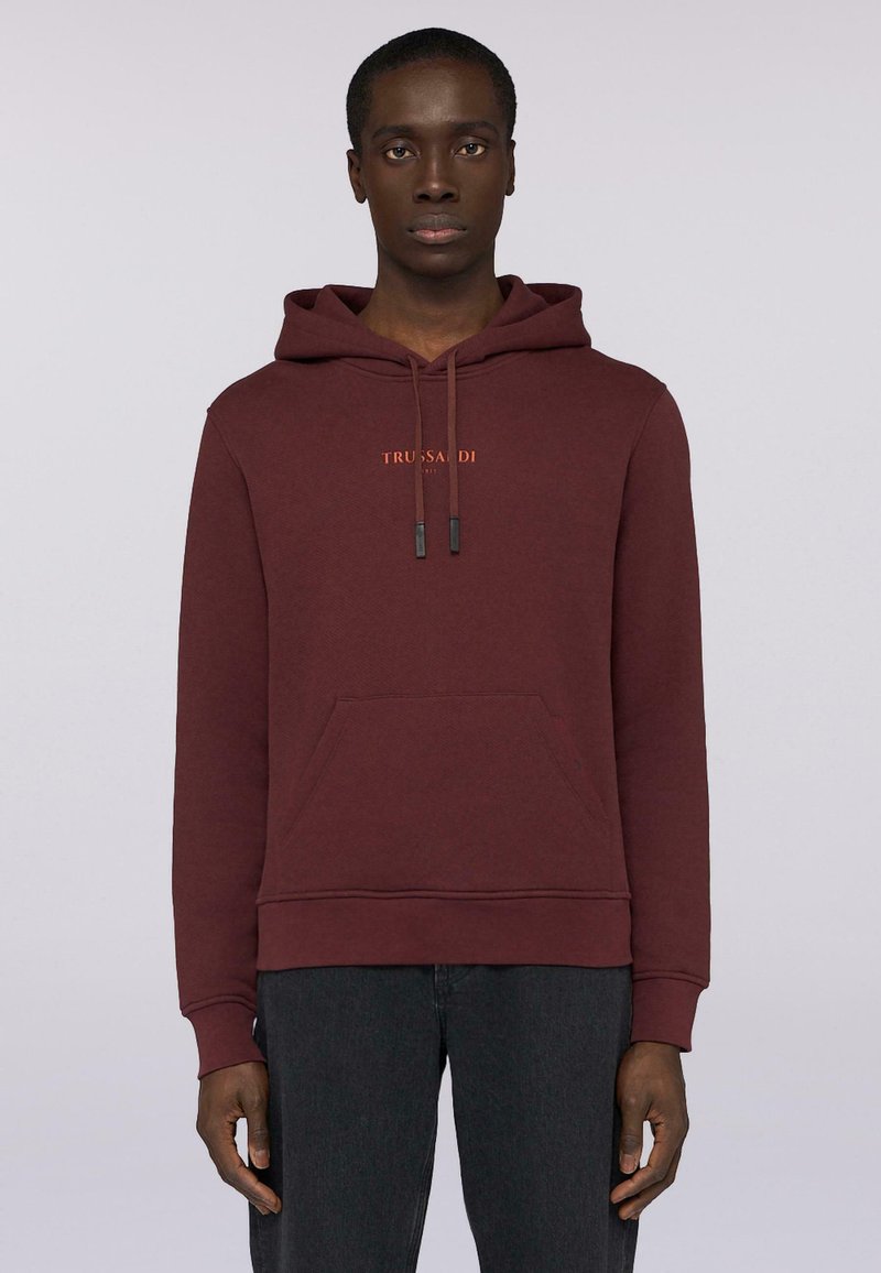 Bordowa bluza z kapturem, wykonana z teksturowanej tkaniny, wyposażona w przednią kieszeń, sznurki oraz haftowane logo w kolorze pomarańczowym. Noszona z ciemnymi dżinsami.