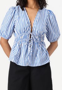 Gestreifte Bluse aus blau-weißem Baumwollstoff, mit tiefem V-Ausschnitt, Puffärmeln, geraffter Taille und Kordelzugdetails. Entworfen mit lockerer Passform.