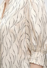 Blouse crème avec motif abstrait linéaire et ponctué noir, tissu texturé, manche bouffante et large poignet froncé sur le bras et l'épaule d’une personne.