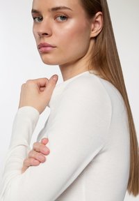 Maglietta bianca a maniche lunghe con un collo alto. Il modello poggia il mento sulla mano, evidenziando una trama liscia e un design aderente.