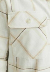 Camisa de cuadros clara en crema, con patrones de check en beige y marrón. Textura suave con un detalle de bolsillo en el pecho con botones.