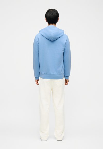 Άτομο που στέκεται γυρισμένο με την πλάτη, φορώντας μια ανοιχτό μπλε μπλούζα με κουκούλα και φαρδιά λευκά παντελόνια, μπροστά σε έναν απλό άσπρο φόντο.